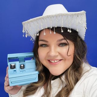 A woman wearing a cowboy hat holding a blue polaroid camera.