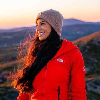 A woman in a red jacket standing on top of a mountain at sunset.