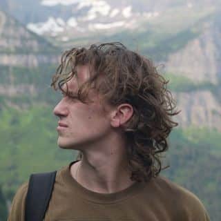 A young man with curly hair standing in front of a mountain.