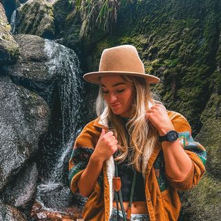 blonde travel influencer wearing an hat and standing before rocks in the nature.