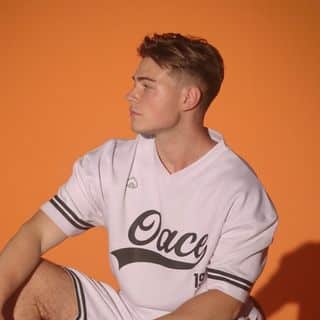 A man in a white baseball jersey sitting on an orange background.