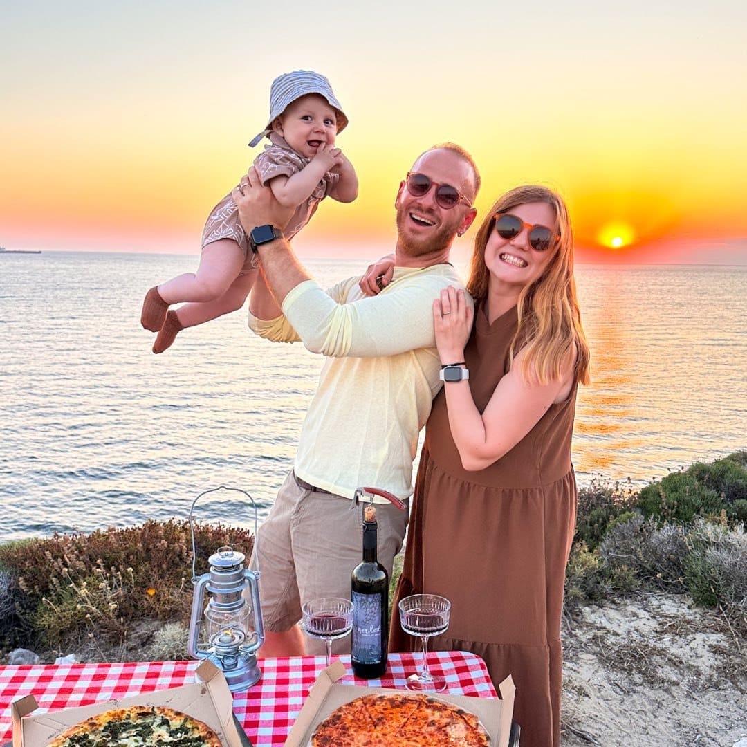 A man and woman holding a baby while eating pizza on the beach.