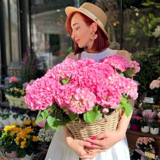 A woman in a hat and a hat holding a basket of pink flowers.