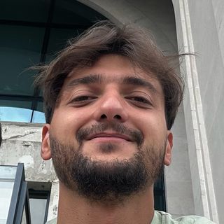 A man with a beard is taking a selfie in front of a building.