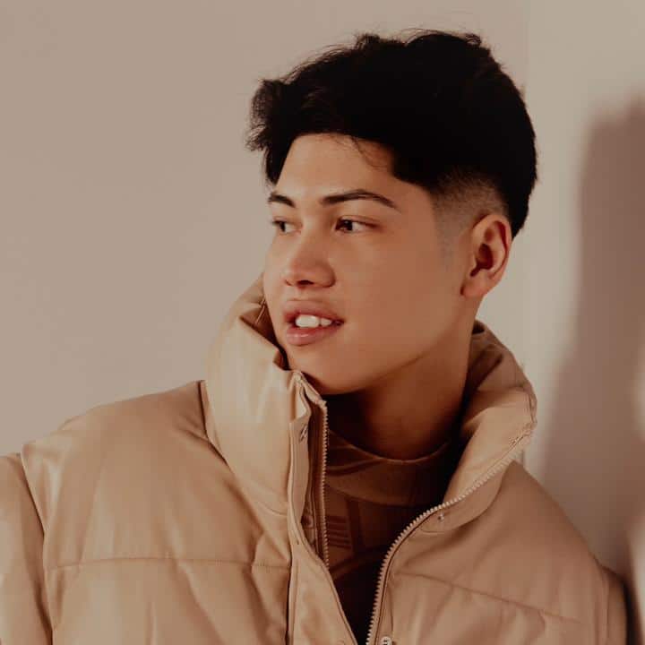 A young man in a beige jacket leaning against a wall.