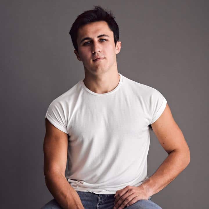 A young man in jeans and a white t - shirt sitting on a gray background.