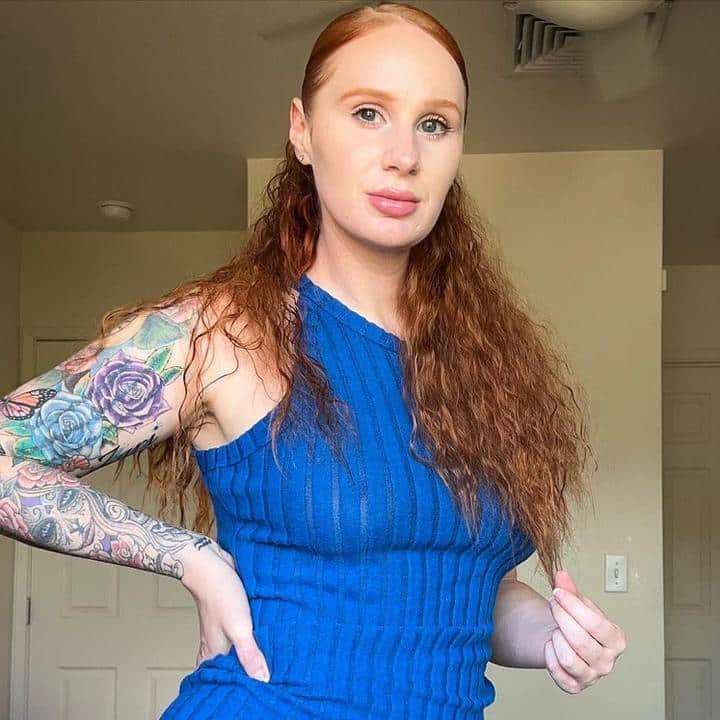 A woman with red hair and tattoos posing in a blue dress.