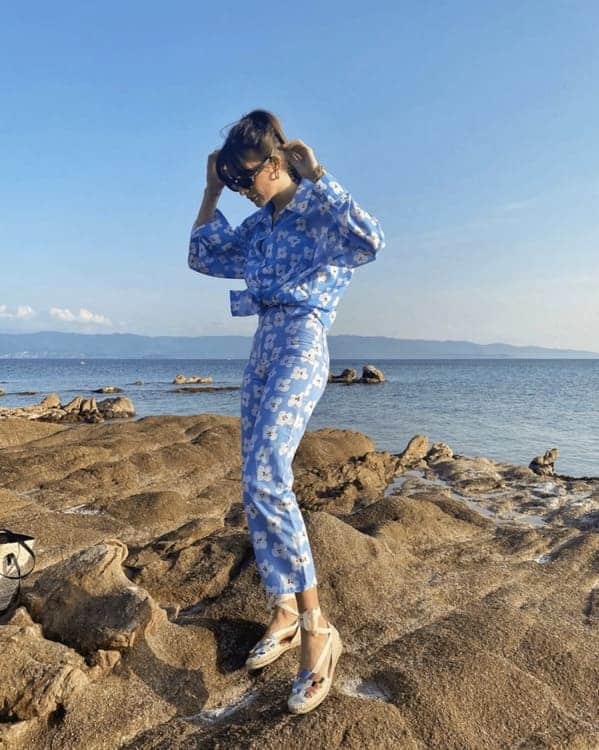 A woman standing on a rocky beach in a blue printed jumpsuit.