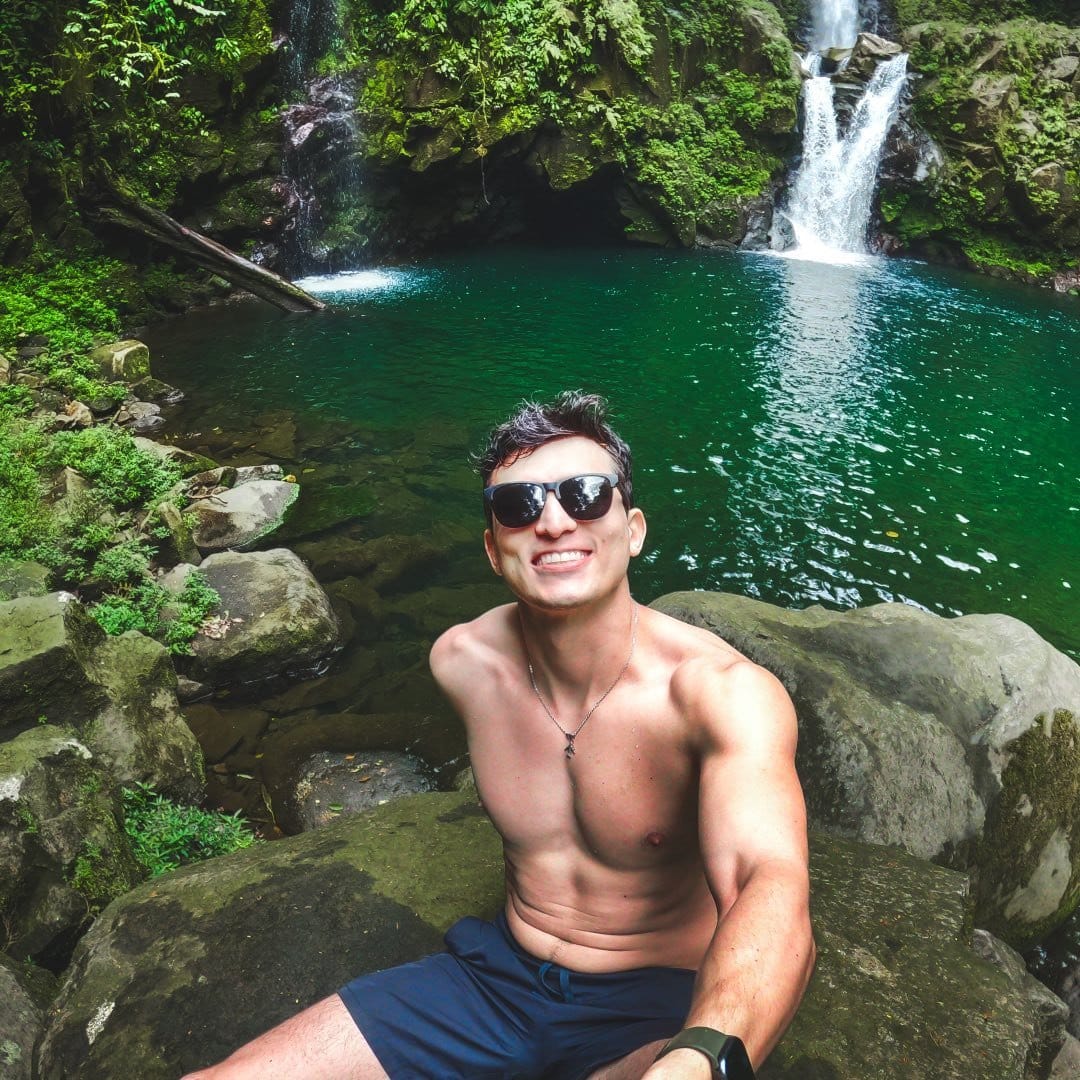 A man in shorts and a t-shirt is taking a selfie in front of a waterfall.