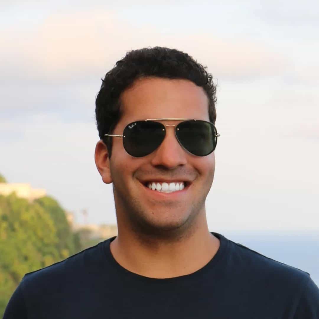 A man in sunglasses smiling in front of the ocean.
