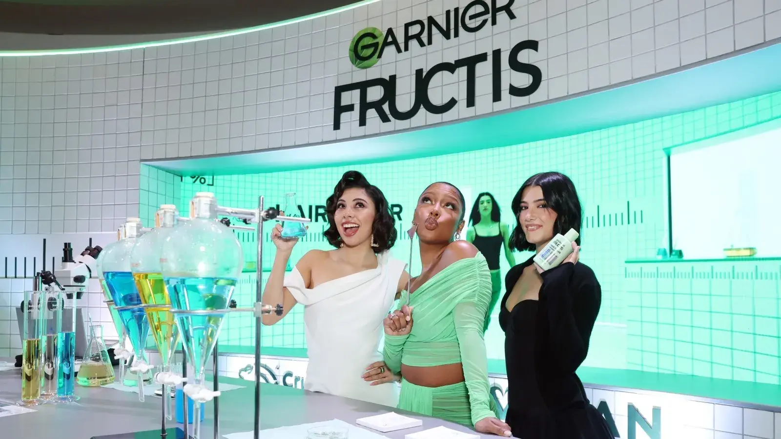 Three women posing with garnier fructis products at a promotional event with a stylized laboratory setup in the background.