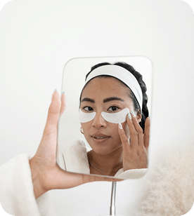 A Woman Wearing A White Headband Applies Under-Eye Patches While Looking At Her Reflection In A Handheld Mirror. A woman wearing a white headband applies under-eye patches while looking at her reflection in a handheld mirror.