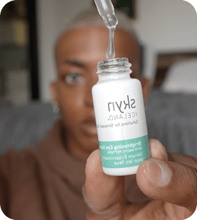 A Person Holds A Dropper Above An Open Bottle Of Skin Iceland Brightening Eye Serum, With The Bottle Label Visible In The Foreground. A person holds a dropper above an open bottle of SKIN ICELAND Brightening Eye Serum, with the bottle label visible in the foreground.