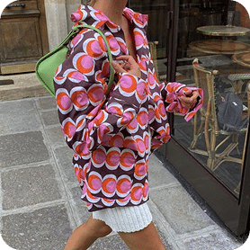 Person Walking On A City Sidewalk Wearing An Oversized, Patterned Shirt With Red And Pink Circles, White Shorts, And Carrying A Small Green Bag On Their Shoulder. Person walking on a city sidewalk wearing an oversized, patterned shirt with red and pink circles, white shorts, and carrying a small green bag on their shoulder.