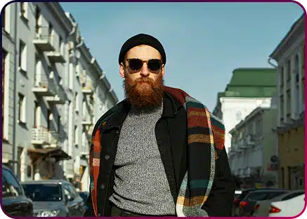 A man with a beard and sunglasses, wearing a beanie and a plaid scarf, stands on a city street lined with buildings and parked cars.