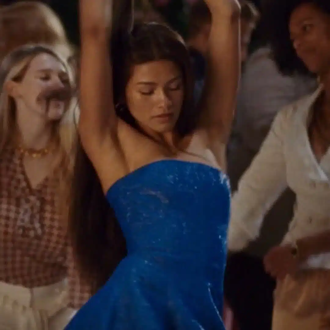 A woman in a blue strapless dress dances with her arms raised, surrounded by other people at a party.
