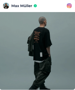 A Person With A Shaved Head Faces Away, Wearing A Black Oversized T-Shirt With Graphics On The Back, Baggy Dark Pants, And Holding A Jacket Over One Shoulder Against A Plain Background. A person with a shaved head faces away, wearing a black oversized t-shirt with graphics on the back, baggy dark pants, and holding a jacket over one shoulder against a plain background.