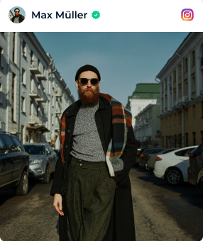 A Man With A Beard Wearing Sunglasses, A Black Coat, And A Scarf Walks Down A City Street Lined With Parked Cars And Buildings. A man with a beard wearing sunglasses, a black coat, and a scarf walks down a city street lined with parked cars and buildings.