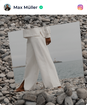 A Person In White Wide-Leg Trousers Stands Barefoot On Pebbles Near A Body Of Water Under An Overcast Sky. A person in white wide-leg trousers stands barefoot on pebbles near a body of water under an overcast sky.