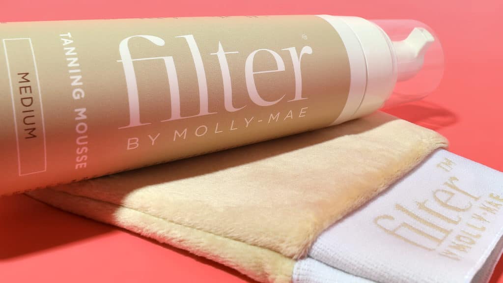Who Is Molly Mae? How She Built A £Multi-Million Brand Empire In 2026 3 A bottle of Filter by Molly Mae tanning mousse in medium shade rests on a Filter-branded tanning mitt against a pink background, reflecting the influencer’s signature glow admired by Love Island fans.