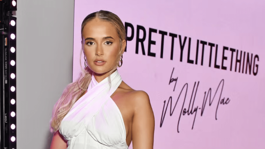 Who Is Molly Mae? How She Built A £Multi-Million Brand Empire In 2026 2 A woman in a white halter dress poses in front of a pink sign that reads "PRETTYLITTLETHING by Molly-Mae." Who is Molly Mae? She’s the influencer and creative behind this stylish collaboration.
