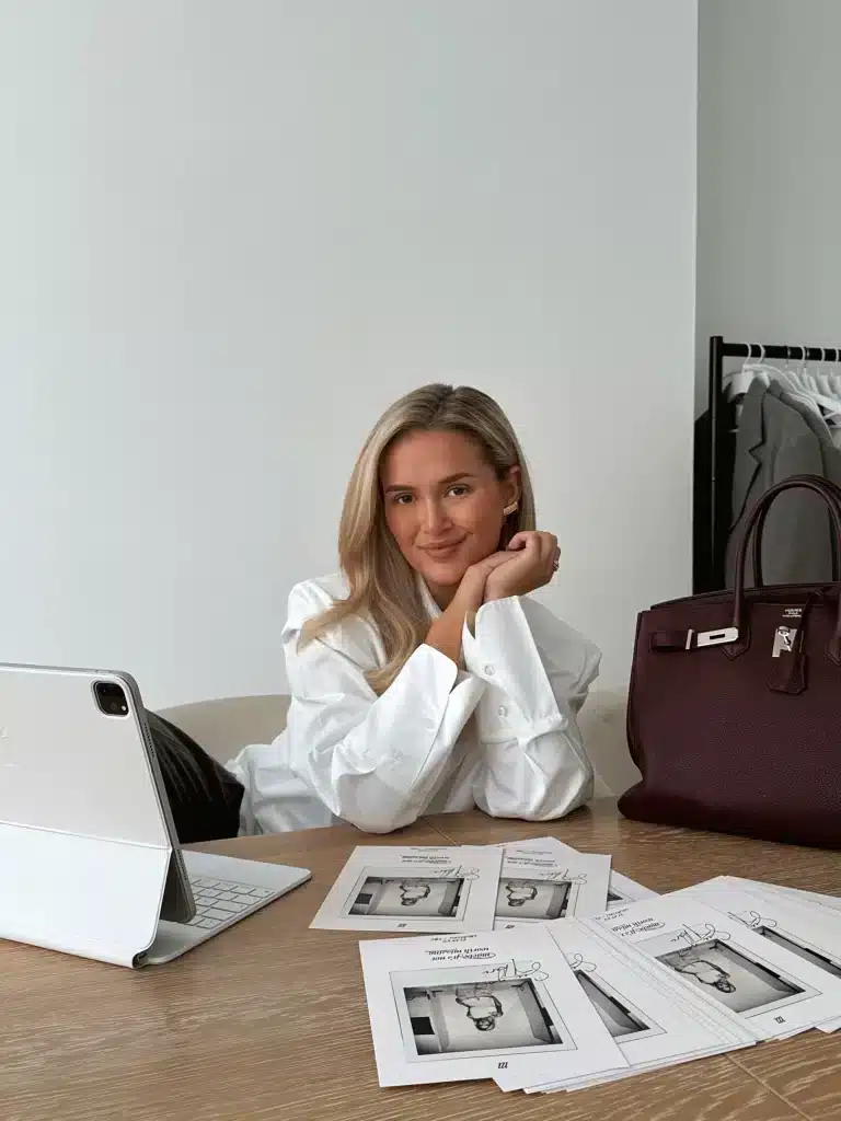 Who Is Molly Mae? How She Built A £Multi-Million Brand Empire In 2026 4 A woman sits at a desk with a tablet, printed documents, and a large handbag. Clothing hangs on a rack in the background. She is looking at the camera and smiling slightly, exuding the style of an influencer like Molly Mae from Love Island.