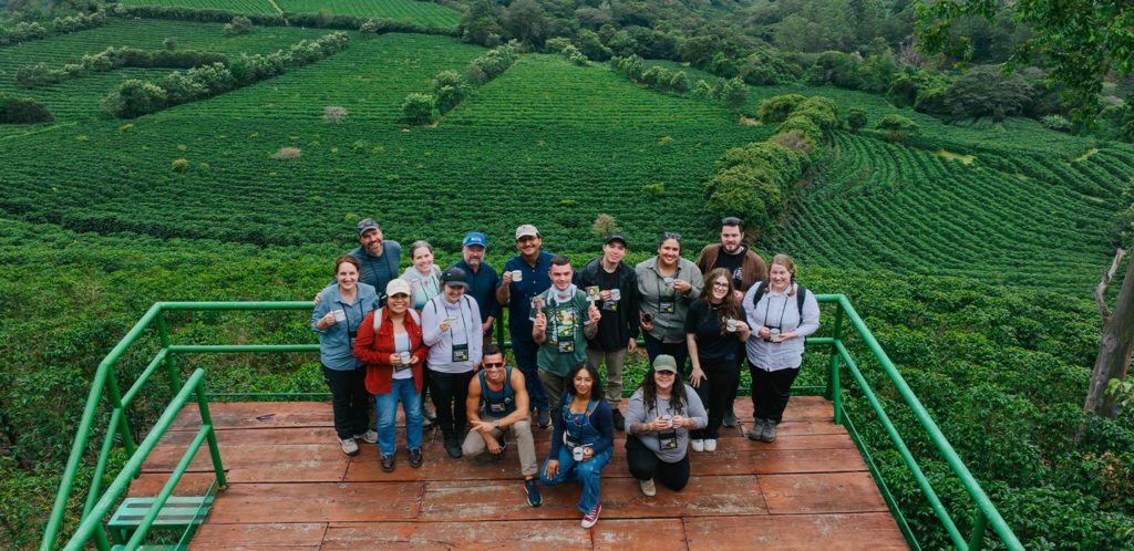 What Is Brand Advocacy? (7 Top Brand Programs Explained) 23 A group of people, united by brand advocacy, poses on a wooden platform overlooking green, terraced fields and dense vegetation in a rural landscape.