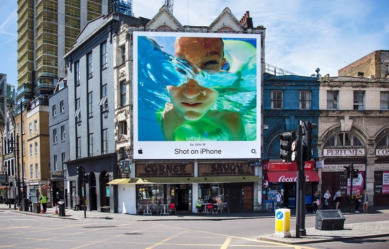What Is Brand Advocacy? (7 Top Brand Programs Explained) 20 A large billboard on a city building displays an underwater photo of a child, with the text "Shot on iPhone," showcasing brand advocacy alongside the Apple logo.